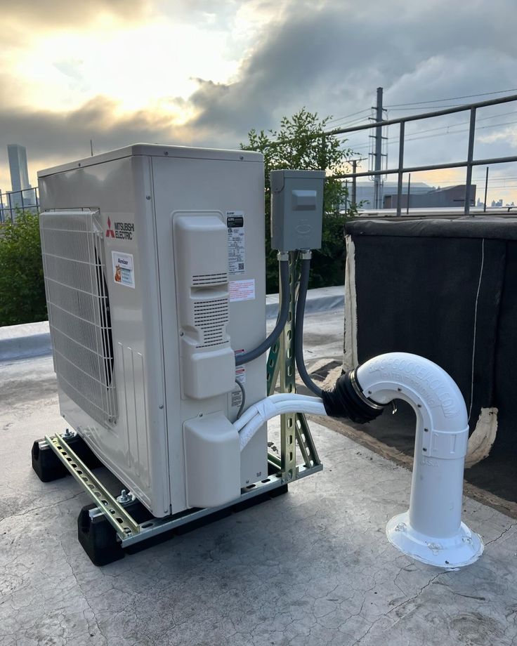 An AiraHome air source heat pump outdoor unit covered in snow with snow on the ground. It is placed against a wall, between some potted plants